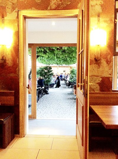 Open doorway from a warmly lit Viennese interior looking out toward a quiet courtyard with tables, greenery, and people in the distance.