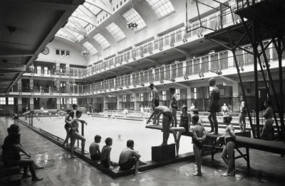 Historical black-and-white photograph of the Amalienbad indoor swimming hall in Vienna, with children and young people gathered beside the pool and on a diving platform beneath a high glazed roof and several levels of changing cabins.