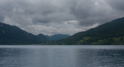 View across the Attersee under dark clouds, with calm lake water in the foreground and a line of villages, wooded slopes and mountains along the opposite shore.