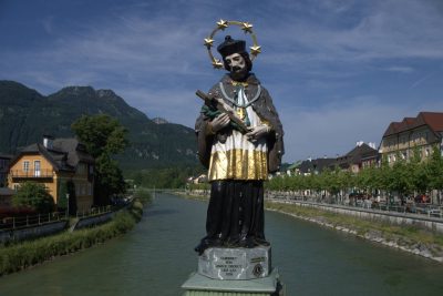 Statue of Saint John of Nepomuk on a bridge in Bad Ischl, standing above the river with town buildings along the banks and forested mountains rising in the background.