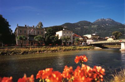View across the river in Bad Ischl towards historic villas and town buildings, with a bridge to the right, bright flowers in the foreground and forested mountains rising behind the town.