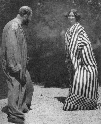 Black-and-white photograph of Emilie Flöge and Gustav Klimt standing outdoors at the Attersee. Flöge wears a long, loose dress with bold vertical stripes and checkerboard bands, while Klimt wears a flowing robe.