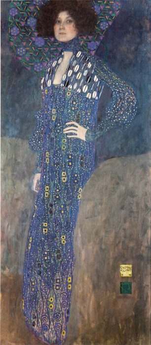 Gustav Klimt’s full-length portrait of Emilie Flöge standing with one hand on her hip, wearing a long blue patterned reform dress with decorative white, gold and violet motifs against a deep blue background.