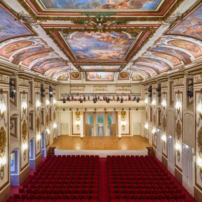 The Haydnsaal at Schloss Esterházy in Eisenstadt, with an elaborately painted ceiling, gilded and floral wall decoration, a stage and rows of red auditorium chairs.