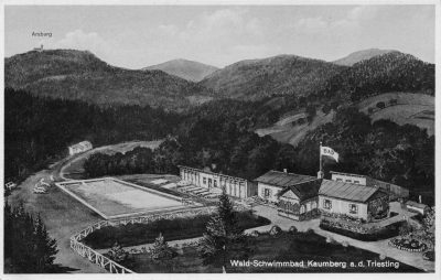 Historical black-and-white postcard of the forest swimming pool at Kaumberg an der Triesting, with a large outdoor pool, wooden bathing and restaurant buildings, landscaped paths and densely wooded hills. The ruins of the Araburg appear on a distant hilltop.