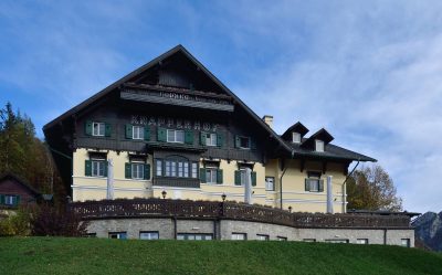 Frontal view of the Knappenhof near Reichenau an der Rax, a large alpine hotel with a pale yellow façade, dark timber upper storeys, green shutters, balconies and a broad terrace beneath a blue sky.