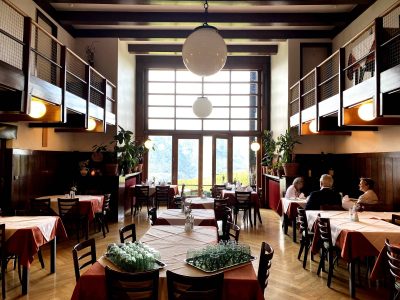 Central interior of Landhaus Khuner, showing a double-height dining room with dark wood beams, upper galleries on both sides, hanging globe lights, large rear windows and tables set beneath a high ceiling.