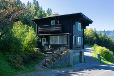 Exterior of Landhaus Khuner near Payerbach, a compact dark timber house with green shutters, a broad overhanging roof, balcony and stone base, set beside a narrow road amid forested mountain scenery.