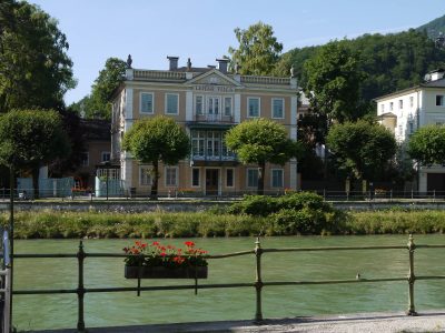 Lehár Villa in Bad Ischl, a pale historicist house with green shutters and a central balcony, seen across the River Traun beneath trees and wooded hills.