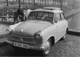 Historical black-and-white photograph of a compact Lloyd car parked on a cobbled road beside a riverside promenade, with two people standing near benches and trees in the background.
