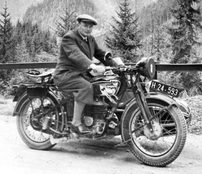 Historical black-and-white photograph of a man in a long coat and flat cap seated on a Puch motorcycle beside a mountain road, with dense conifer forest and steep alpine slopes behind him.