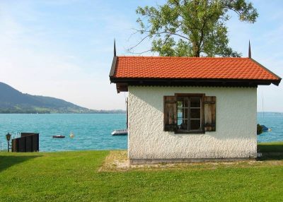 Small white lakeside hut with a red tiled roof and wooden shutters at Steinbach am Attersee, standing on a grassy shore beside the blue water with low hills in the background.