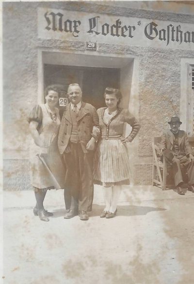Historical photograph of two women and a man standing outside Max Locker’s Gasthaus, numbered 29, while another man wearing a hat sits on a chair beside the entrance.