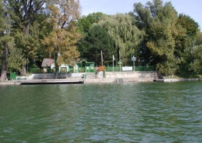 View from the Alte Donau towards the Naturfreunde bathing dock, with wooden swimming platforms, steps leading into the water, green railings and small buildings beneath dense trees.