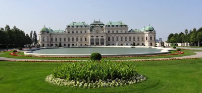 The Upper Belvedere in Vienna seen across its reflecting pool and formal gardens, with a long white Baroque façade, green copper roofs, clipped lawns and flowerbeds.