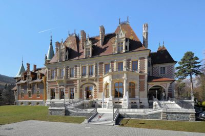 Exterior view of Schloss Rothschild in Reichenau an der Rax, a large historic villa-like residence with ornate façades, steep patterned roofs, towers, terraces and broad entrance steps, set against a clear blue sky.