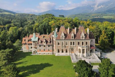 Aerial view of Schloss Rothschild in Reichenau an der Rax, an ornate historicist residence with patterned roofs, towers and terraces, surrounded by lawns, dense woodland and mountain slopes.