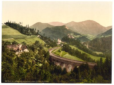 Historic coloured view of the Semmering Railway winding through the mountain landscape at the Jägergraben, with a curving rail line crossing green hills and alpine slopes, and scattered buildings in the valley.