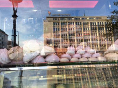 Vienna street architecture reflected in a confectionery window behind rows of white and pink meringues, with interior lights and the city layered across the glass.