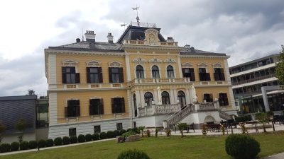 Garden façade of Villa Seilern in Bad Ischl, a large pale yellow historicist villa with dark shutters, ornate white architectural decoration, arched windows, a central terrace and broad stone steps beneath a cloudy sky.