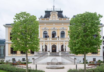 Garden façade of Villa Seilern in Bad Ischl, a pale yellow historicist villa with ornate white decoration, arched windows, a central balcony, broad stone steps, a fountain and two large trees.